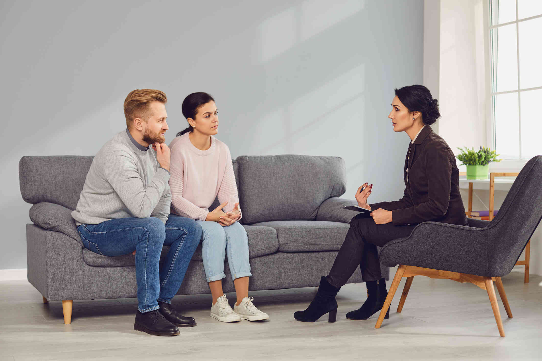 A couple seated on a couch listens intently to a therapist seated across from them, both appearing focused and serious.